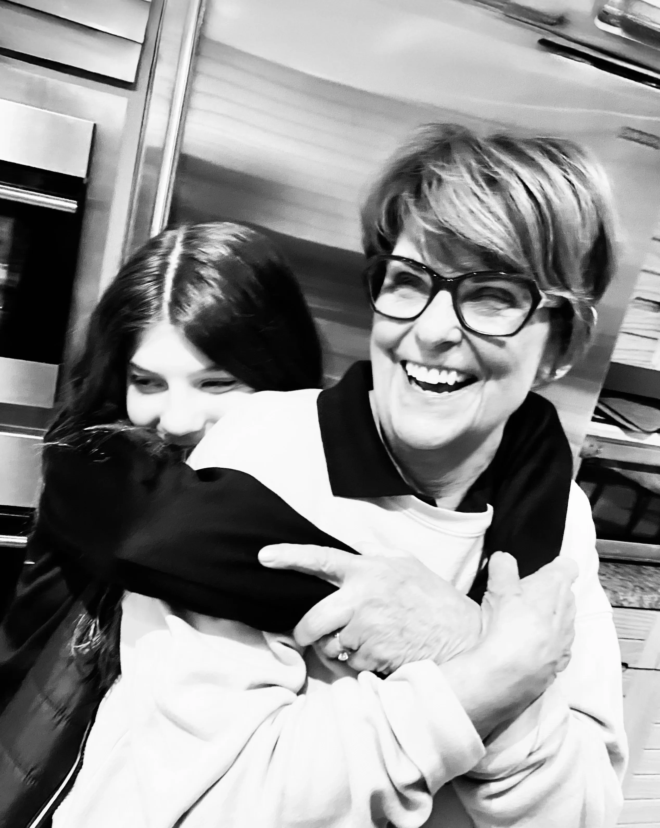 Smiling older woman wearing glasses being hugged from behind by a younger person with long hair.